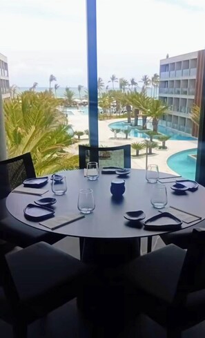 A dining table with chairs, a view of a pool, palm trees, and a building in the background. - Apartment Porto 2 Live Resort Muro Alto - Porto de Galinhas (Ipojuca)