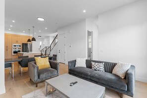 A modern living room with a grey sofa, a coffee table, and a dining area with a kitchen in the background. - Modern Luxury Home | 3 Min to University, Stadium & Major Hospitals (Calgary)