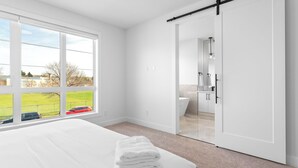A bedroom with a bed, a window with a view, a sliding door to a bathroom, and a white color scheme. - Modern Luxury Home | 3 Min to University, Stadium & Major Hospitals (Calgary)
