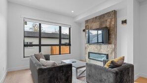 A modern living room with a sofa, armchair, and a flat-screen TV mounted on a stone wall. - Modern Luxury Home | 3 Min to University, Stadium & Major Hospitals (Calgary)
