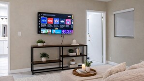 Un salon avec un téléviseur à écran plat fixé au mur, une console en bois agrémentée d’objets décoratifs, un tapis blanc et moelleux, et une porte sur le côté droit.