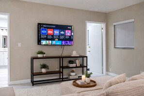 Ein Wohnzimmer mit einem an der Wand befestigten Flachbildfernseher, einem Holzkonsolentisch mit Dekorationen, einem weißen, flauschigen Teppich und einer Tür rechts.