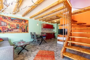 A living room with a sofa, a dining table, and chairs. There is a kitchen area with a red cabinet and a TV. A wooden staircase is visible. - Mexican-themed Holiday Homes (Ustka)