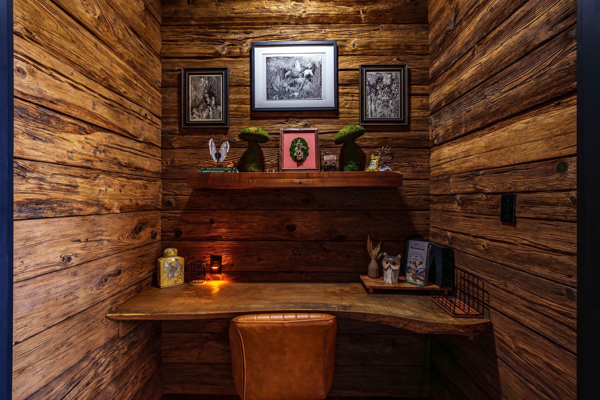 A rustic wooden room with a wooden desk, framed pictures, and a small shelf with decorative items.
