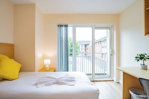 A hotel room with a bed, a desk, a chair, a lamp, a potted plant, and a sliding glass door leading to an outdoor area.