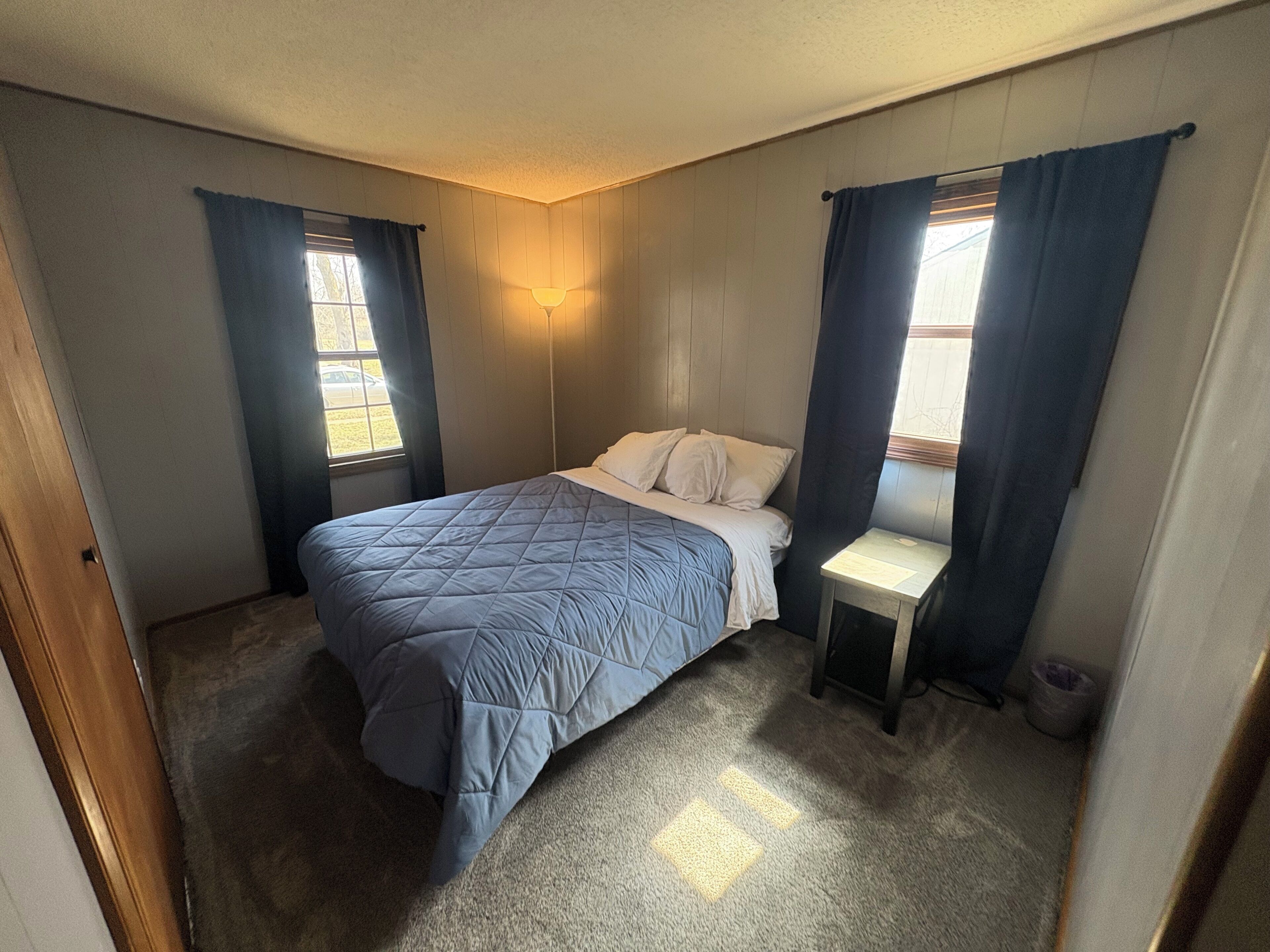A bedroom with a bed, a nightstand, a window with curtains, and a wall-mounted light fixture.