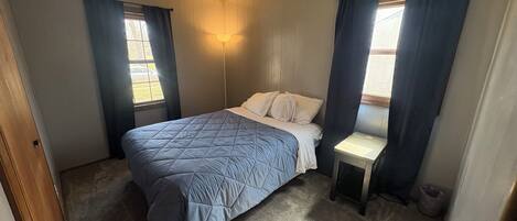 A bedroom with a bed, a nightstand, a window with curtains, and a wall-mounted light fixture.