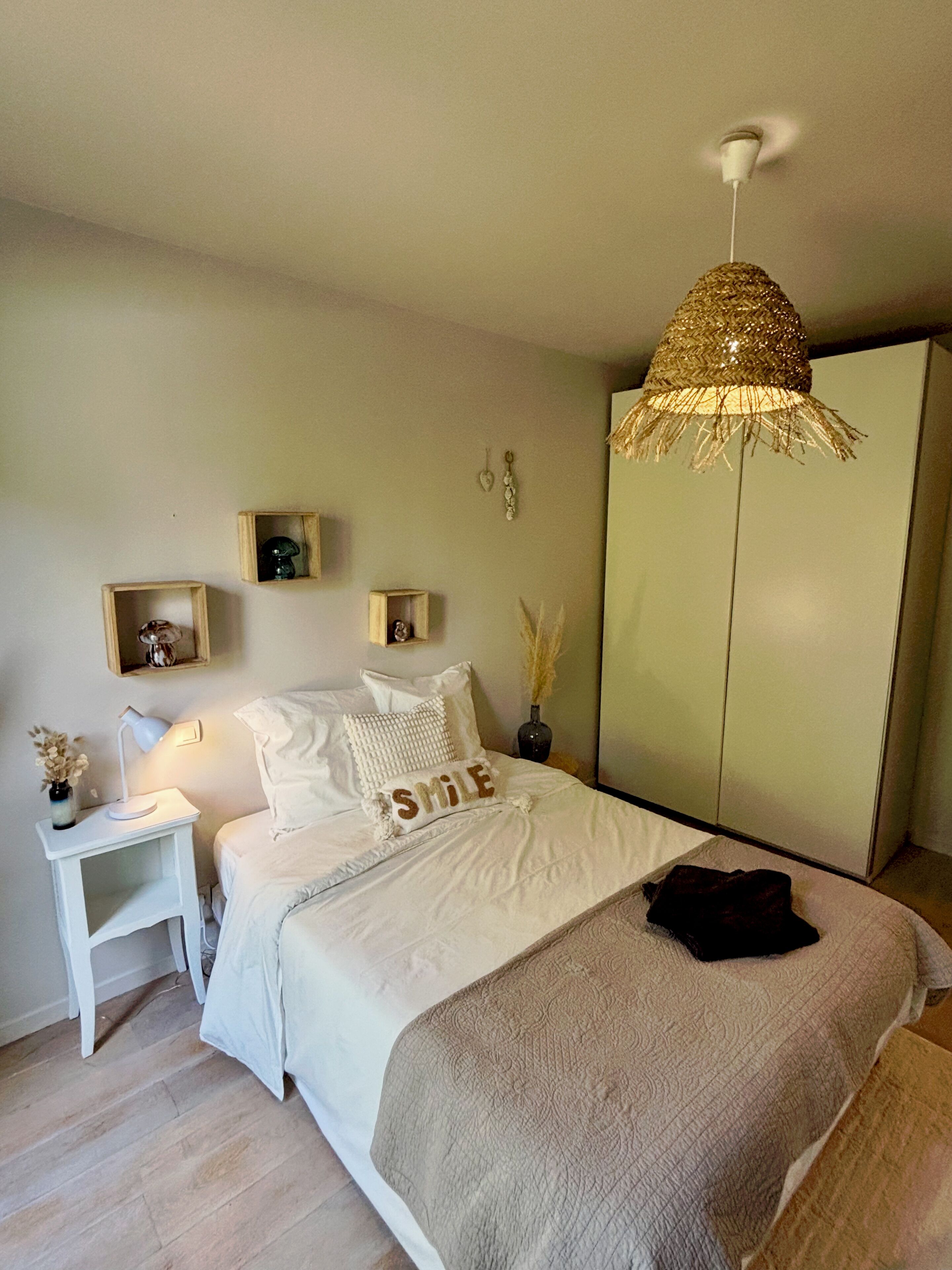 A bedroom with a bed, bedside table, lamp, decorative pillows, a wardrobe, and a hanging lamp.