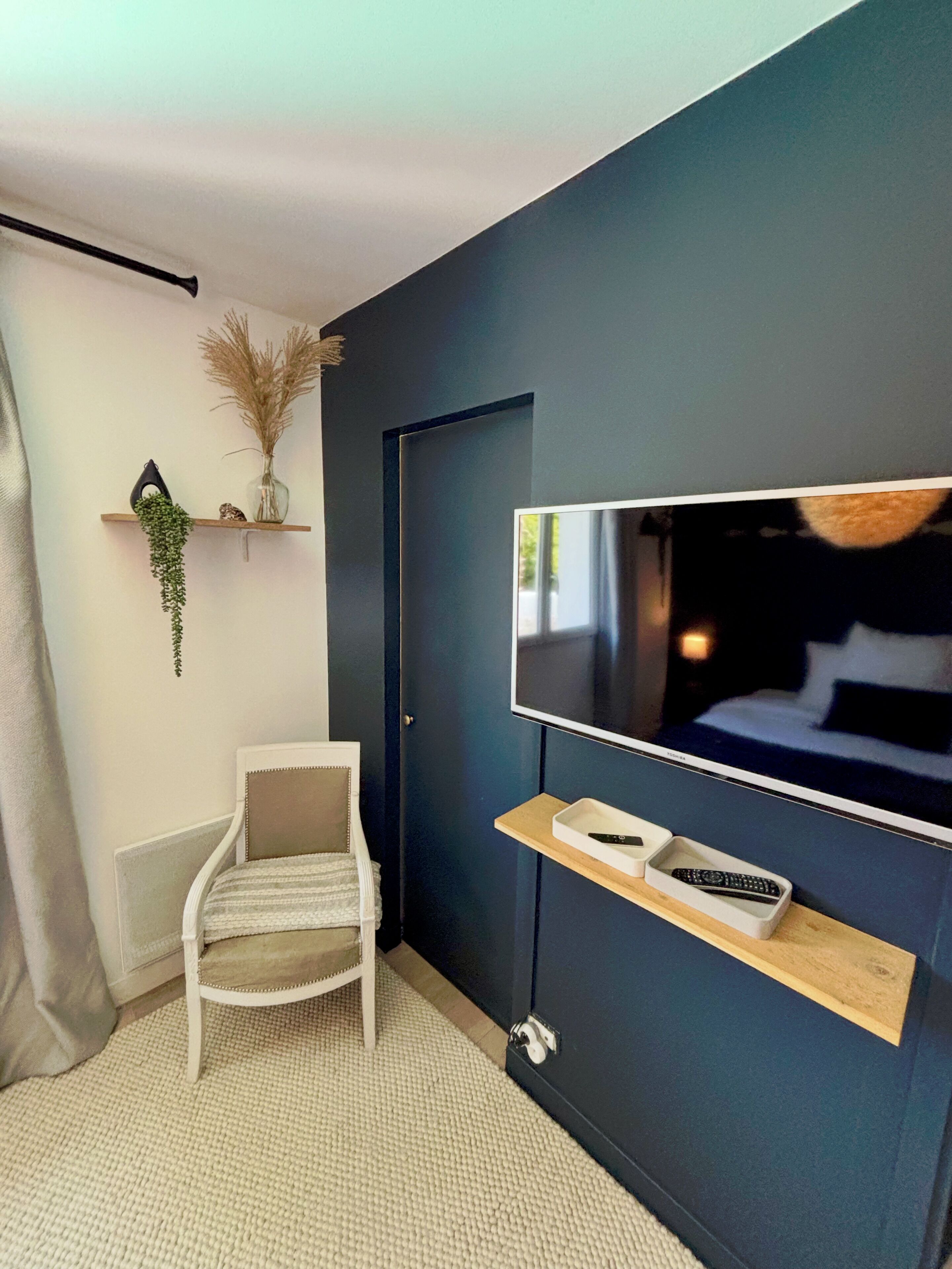 Una habitación moderna con una pared azul oscuro, un televisor montado, una silla blanca, un estante de madera con mandos a distancia y una planta colgante.