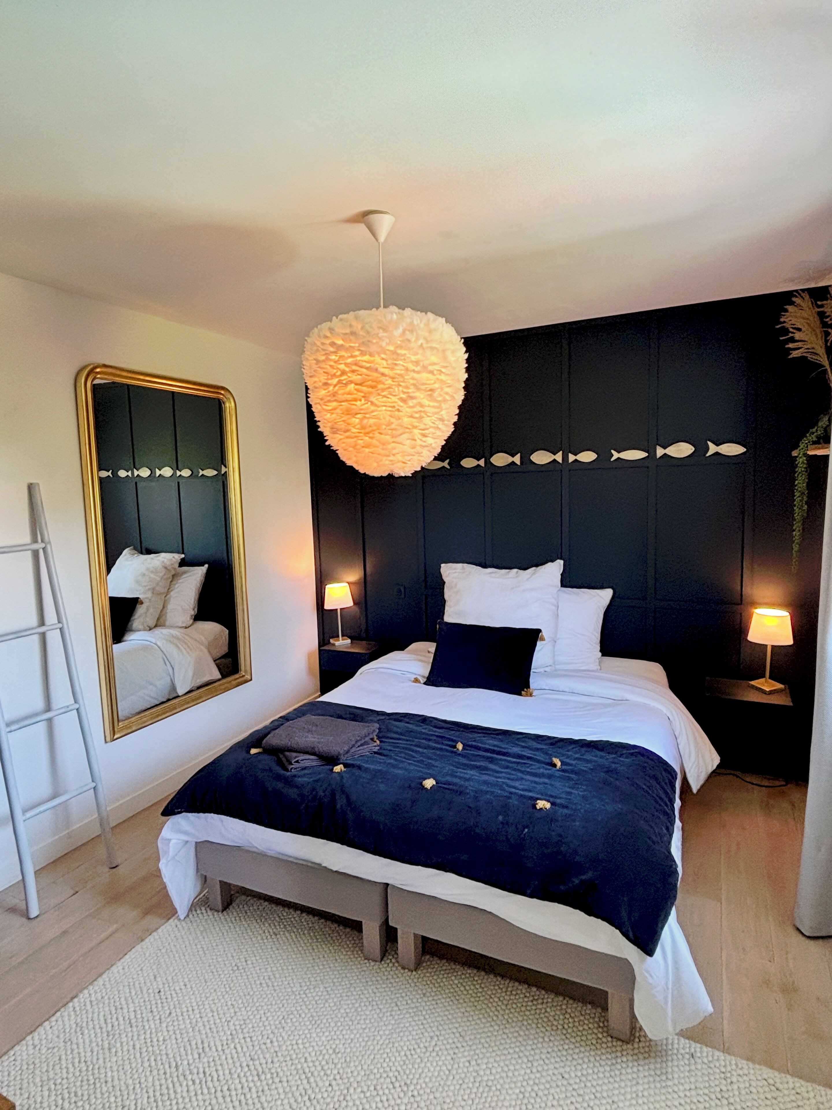 A bedroom with a bed, bedside tables, a mirror, a hanging light fixture, and a ladder.