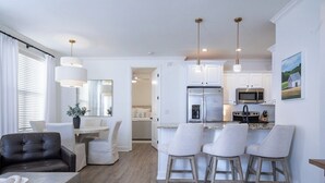 A modern kitchen with white cabinets, a stainless steel refrigerator, and a microwave. There are four bar stools and a dining table with chairs. - The Tanglewood, 3BR by Velvet Ditch Villas (Oxford)
