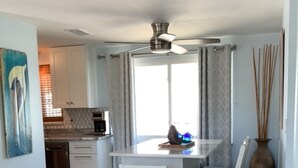 A dining area with a white table and chairs, a ceiling fan, a large window with curtains, and a kitchen in the background.