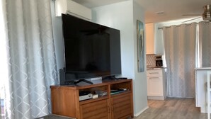 A living room with a flat-screen TV on a wooden stand, a kitchen in the background, and a dining area with a table and chairs.