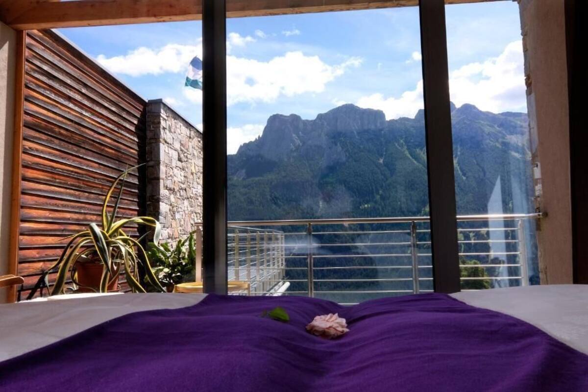 A room with a bed, a balcony with a view of mountains, and a wooden wall.