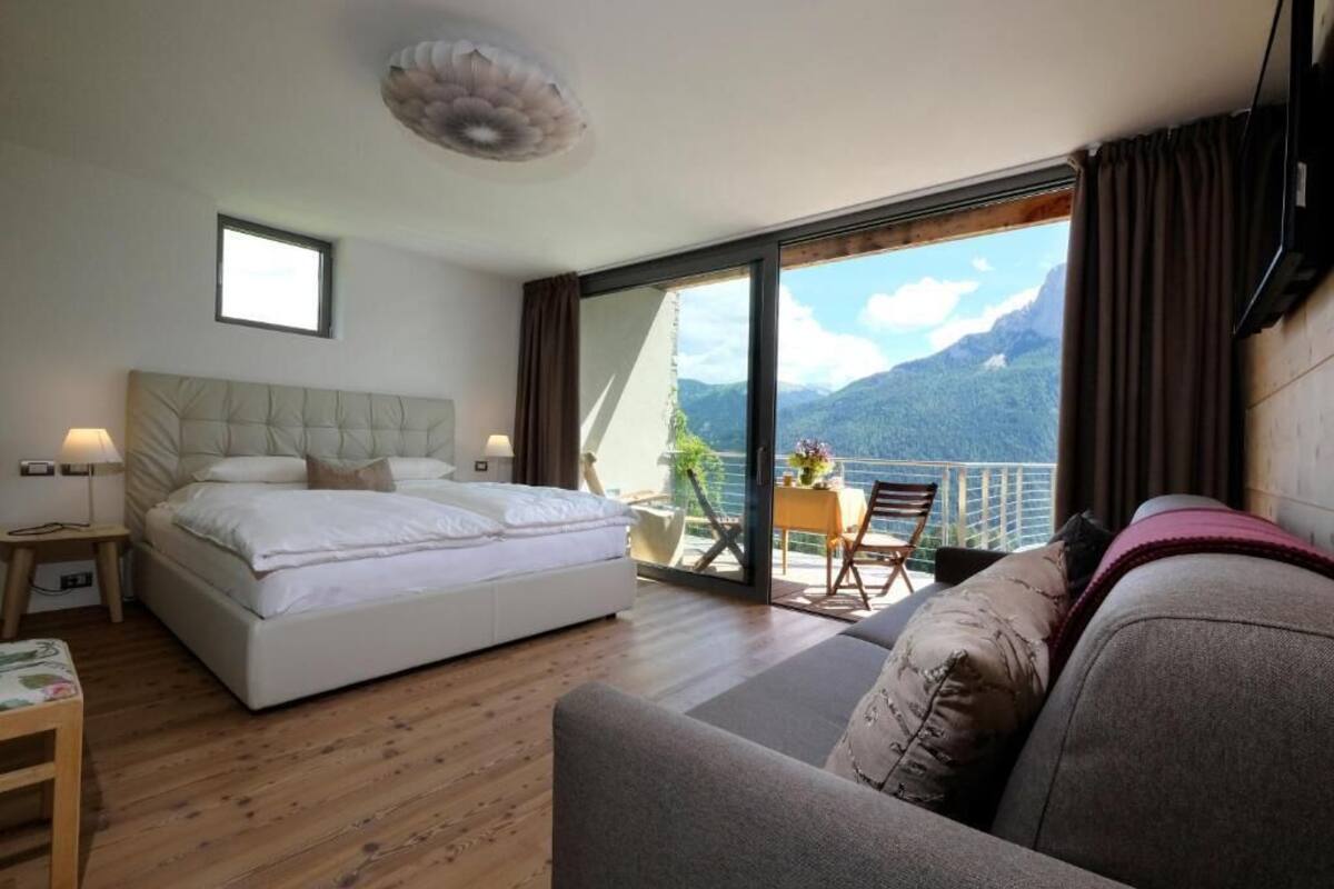 A bedroom with a bed, a sofa, a table with chairs, and a balcony with a mountain view.