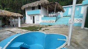 Una piscina azul con borde de piedra, una estructura con techo de paja y un edificio de paredes azules y blancas.