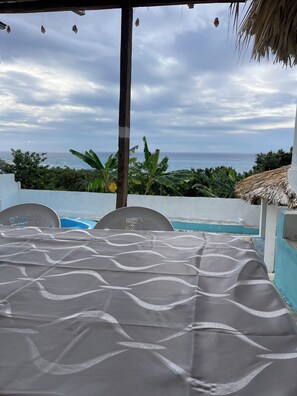 Un espace extérieur aménagé, abrité et doté de sièges, offrant une vue sur l’océan, une piscine et une végétation tropicale.