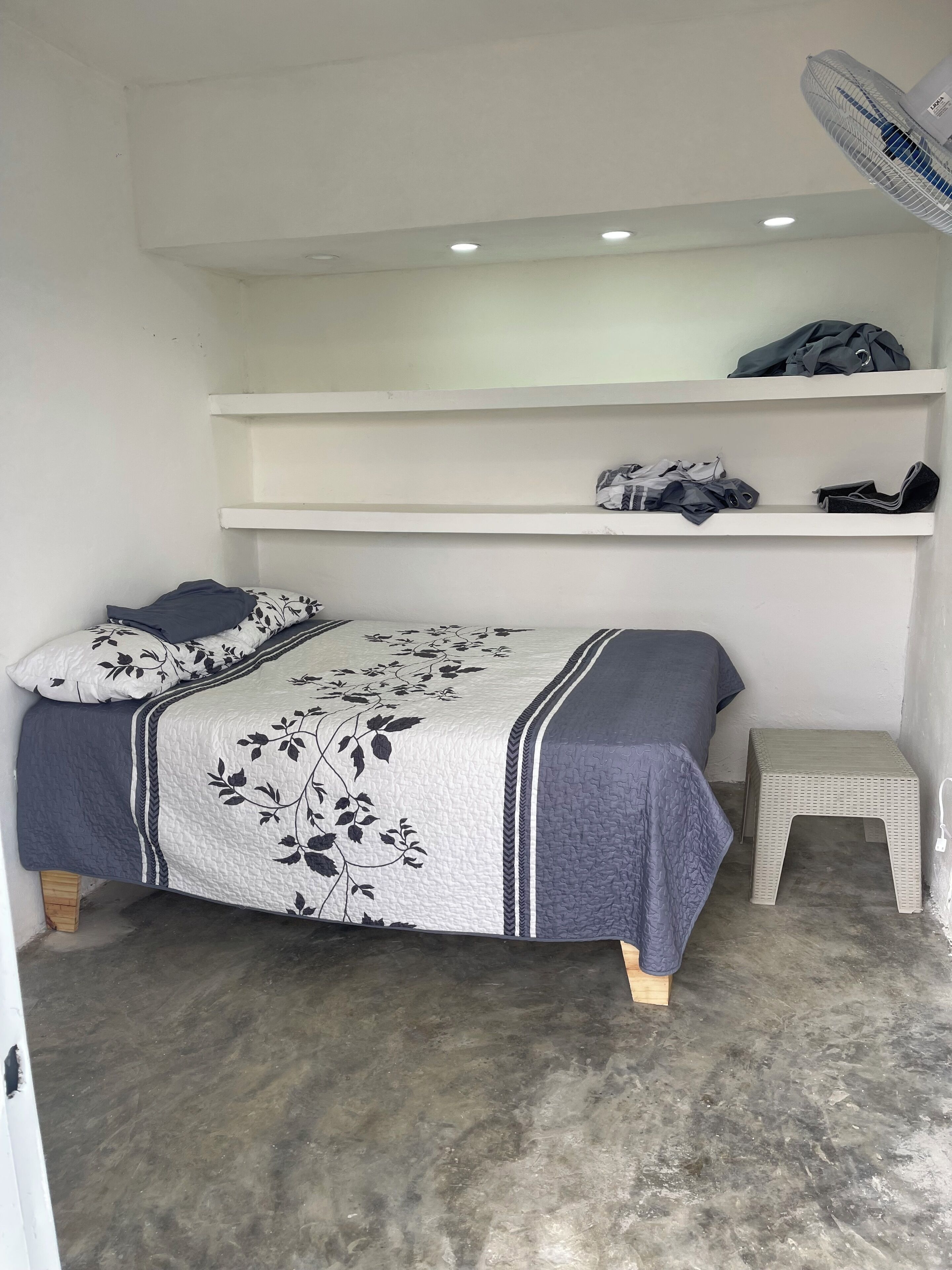Une petite chambre avec un lit simple, recouverte d’une courtepointe à motifs floraux gris et blancs, agrémentée d’un oreiller gris, d’une couverture grise et d’un petit tabouret. Un ventilateur de plafond complète l’ensemble.