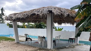 Un área de estar al aire libre con techo de paja, sillas de plástico blancas y una piscina al fondo.