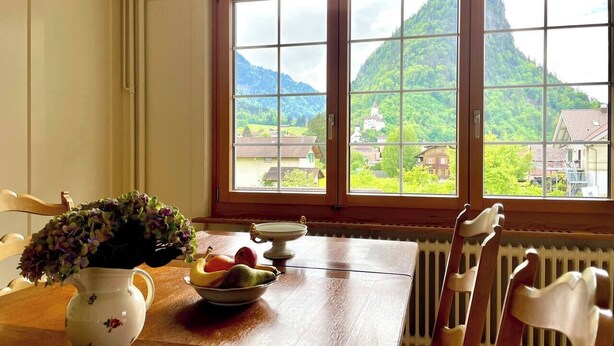 Uma mesa de jantar com um vaso de flores, uma tigela de frutas e uma janela com vista para montanhas e casas.