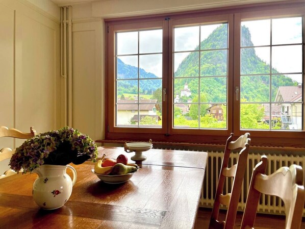 A dining table with a vase of flowers, a bowl of fruit, and a window with a view of a mountain and houses. - Hidden Gem close to Interlaken and Hiking (Wimmis)