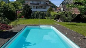 Et rektangulært pool med en træterrasse foran, omgivet af frodig vegetation og et bjerg i baggrunden.