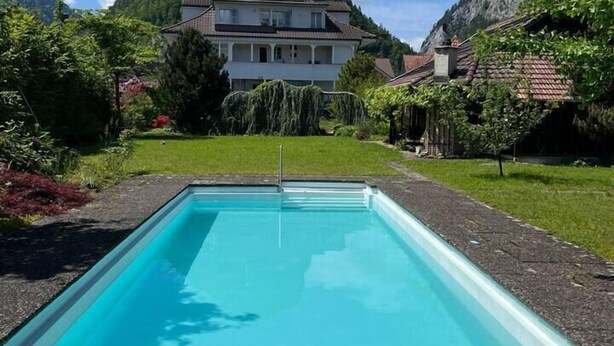 A rectangular swimming pool with a wooden deck in front, surrounded by lush greenery and a mountain backdrop. - Hidden Gem close to Interlaken and Hiking (Wimmis)