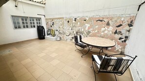 Eine Terrasse mit gefliestem Boden, einem runden Tisch mit Stühlen, einer schwarzen Mülltonne und einem Fenster mit Blick.