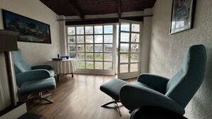 Ein Zimmer mit zwei türkisfarbenen Sesseln, einem kleinen Tisch mit einer blauen Schale und einem großen Fenster mit Blick ins Freie.
