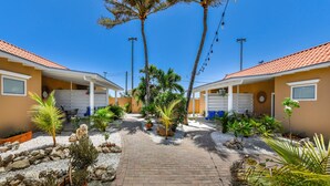 A pathway leading to a row of two-story buildings with a tiled roof, surrounded by various potted plants and a gravel area. - Walts Aruba (Noord)