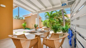A covered patio with a table set for two, surrounded by wicker chairs, a small plant, and a view of a residential area with palm trees. - Walts Aruba (Noord)