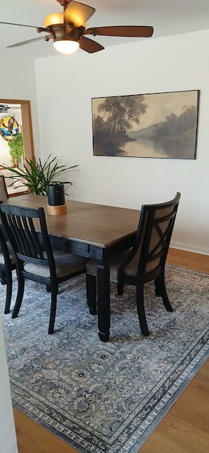 A dining area with a wooden table, black chairs, a patterned rug, a ceiling fan, and a painting on the wall.