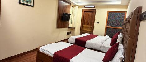 A hotel room with a wooden floor, a bed with white and red bedding, a ceiling fan, an air conditioning unit, a framed picture on the wall, and a wooden door.