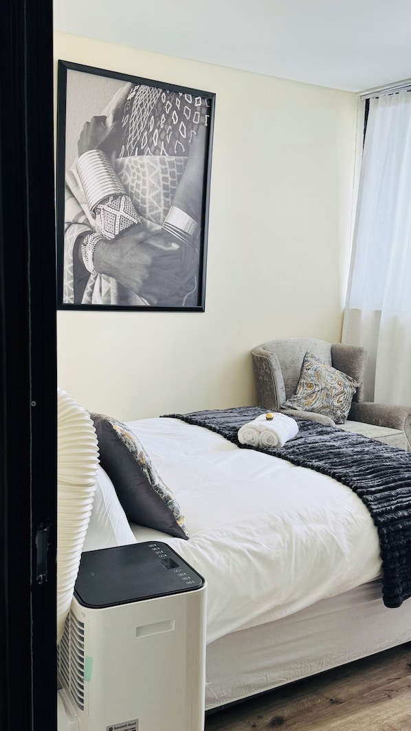 Une chambre à coucher comprenant un lit, une table de chevet, un radiateur, un tableau accroché au mur et une fenêtre avec des rideaux.