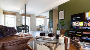 Una sala de estar con un sofá de cuero, una mesa de centro de vidrio, copas de vino, una vela y un televisor mostrando un programa de cocina.
