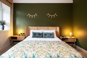 A bedroom with a large bed, two bedside tables, a window with a plant, and a wall with two wooden eye shapes. - Riverside Mill - Luxury Cottages (Kendal)