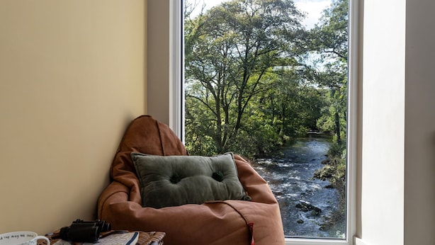 Una habitación con un ventanal grande con vista a un río, un sillón de mimbre, una mesita de mimbre con un libro y una taza, y una cámara.
