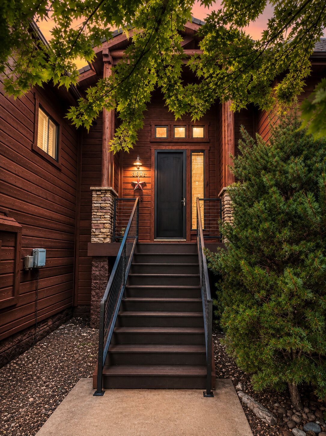 A wooden house with a staircase leading to a dark door, surrounded by trees and a stone wall.