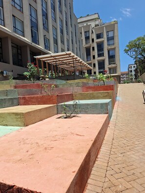A multi-story building with a covered outdoor seating area and a landscaped terrace. - Modern 1-bedroom apartment in tranquil Nairobi - Walk to Everything (Nairobi)