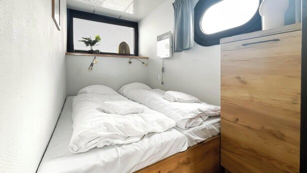 A small, neatly made bed with white bedding, a window with blinds, a wall-mounted air conditioner, and a wooden cabinet. - Boathouse With Lake View Terrace (Uitgeest)
