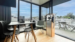A modern room with a large window, a table with chairs, a small wooden cabinet, and a view of a marina. - Boathouse With Lake View Terrace (Uitgeest)