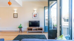 Een woonkamer met een flatscreen televisie, een houten dressoir en een groot raam met uitzicht op gebouwen.