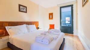 Een hotelkamer met een groot bed, een houten hoofdbord, een lijst met een foto aan de muur, een deur met een raam en een klein tafeltje met een lamp.