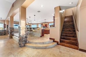 A spacious living area with a stone fireplace, a wooden staircase, and a kitchen in the background.