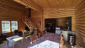 A log cabin interior with a dining area, a kitchen, a staircase, and a view through a window.