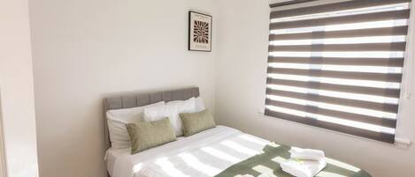 Una cama bien hecha con una manta verde y toallas, un cabecero gris y una ventana con persianas.