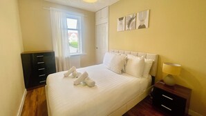En pænt redt seng med puder og håndklæder, et natbord med lampe, en kommode og et vindue med gardiner.