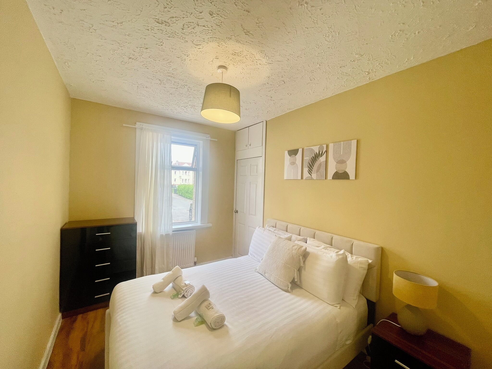A bedroom with a bed, a window, a radiator, a dresser, and wall art.
