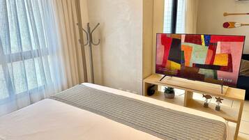 A hotel room with a bed, a TV on a wooden stand, a window with curtains, and a coat rack.