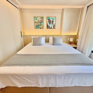 A hotel room with a large bed, two paintings on the wall, a nightstand with a lamp, and a window with curtains. - Loft em Ponta Negra com vista para o mar (Natal)